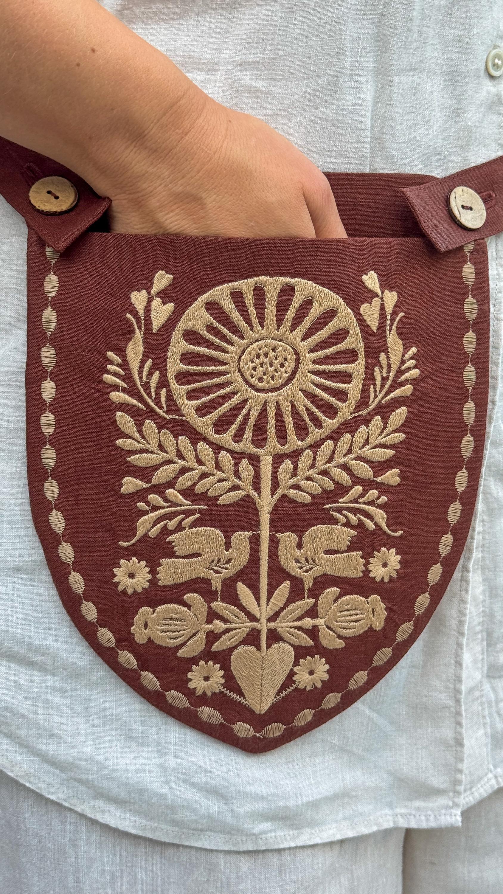 A close-up of a hand holding a burgundy shield-shaped fabric pouch with beige embroidered floral and bird motifs, worn over a white linen shirt.
