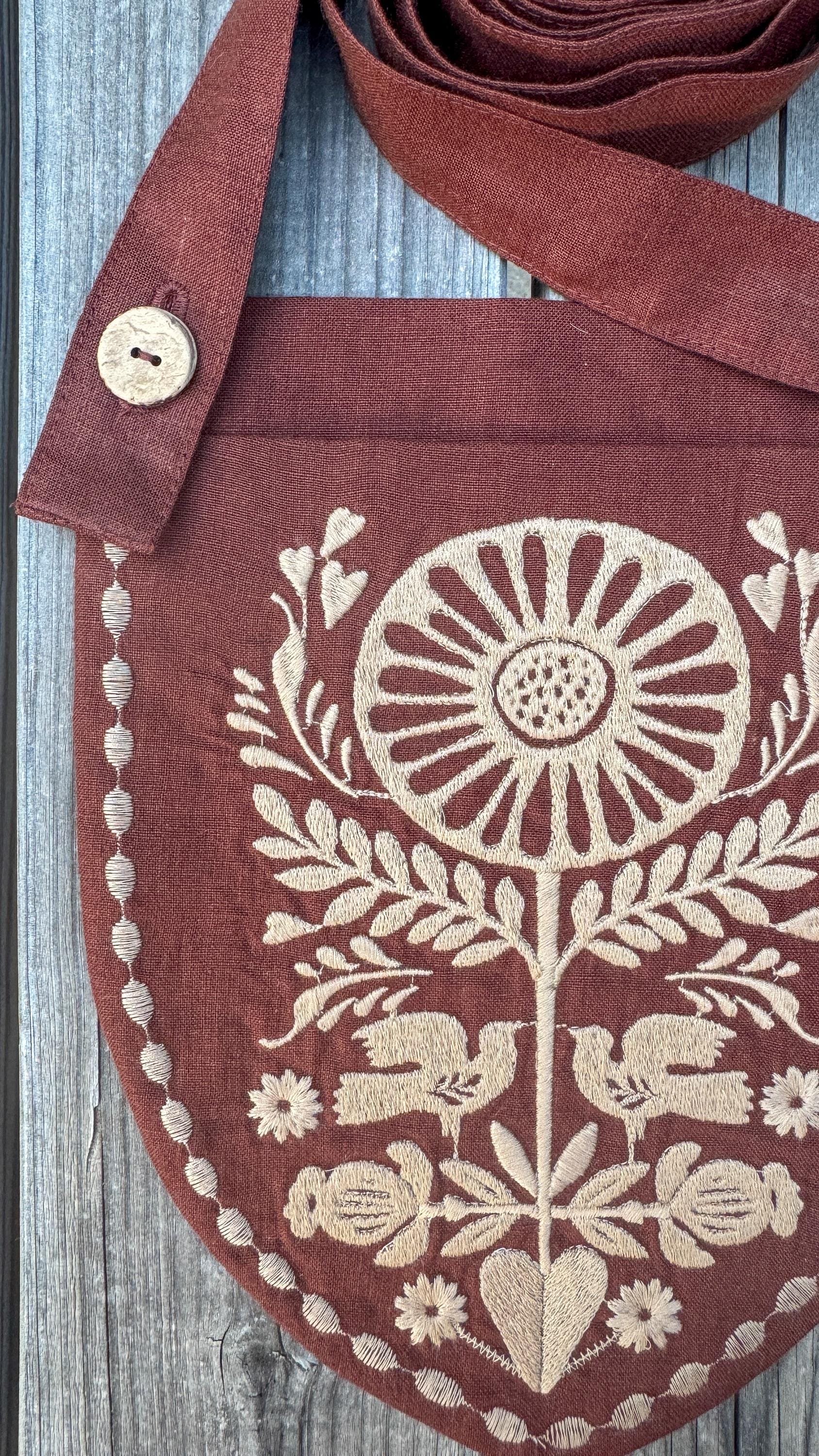 A rust-brown linen crossbody bag with cream-colored embroidery of a sunflower, leaves, birds, and berries, shown on a weathered gray wood background.
