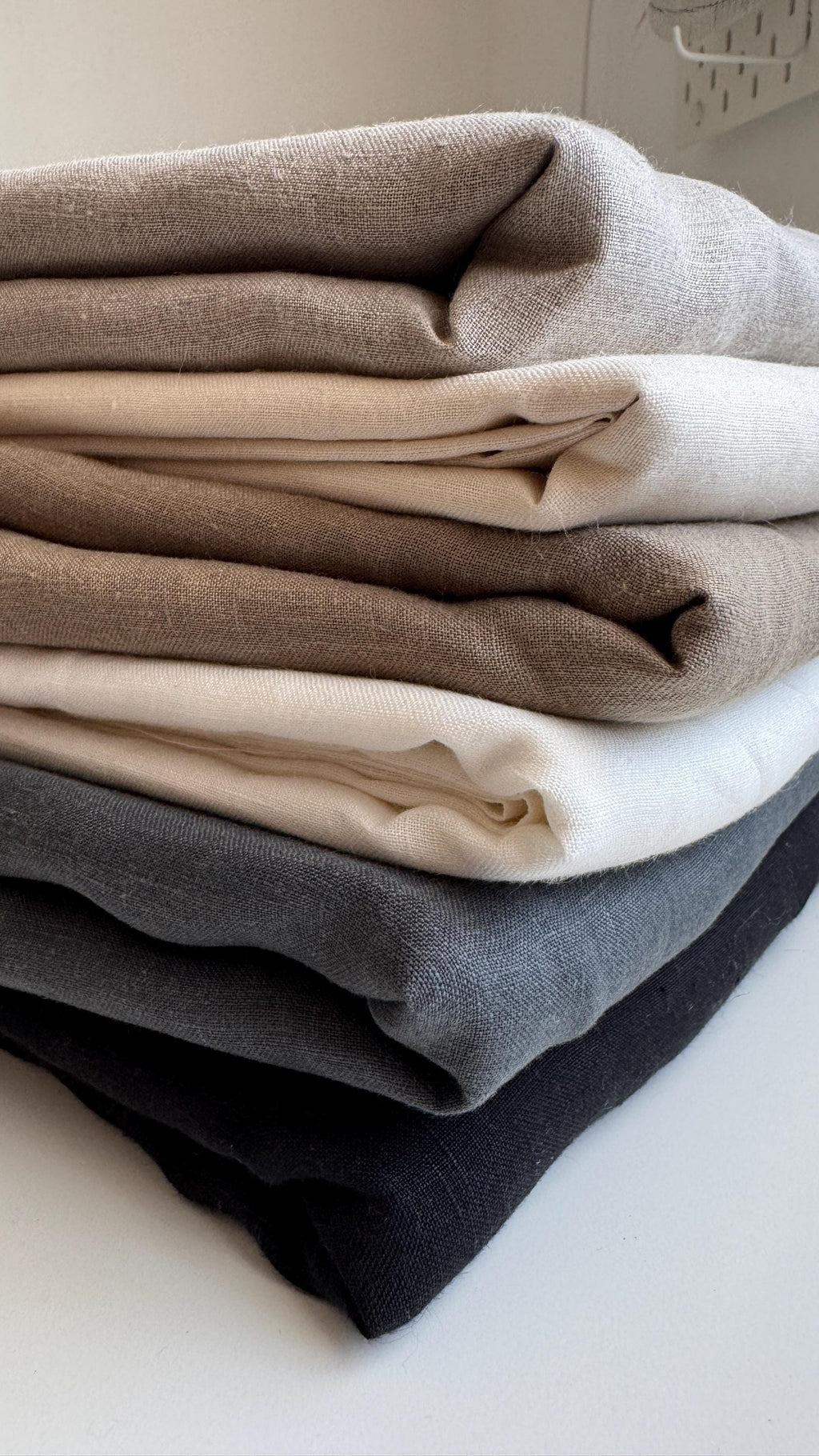 A stack of folded textured fabric pieces in neutral tones, including taupe, cream, light brown, white, charcoal gray, and black, photographed on a light surface.