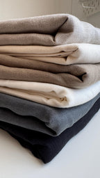 A stack of folded textured fabric pieces in neutral tones, including taupe, cream, light brown, white, charcoal gray, and black, photographed on a light surface.