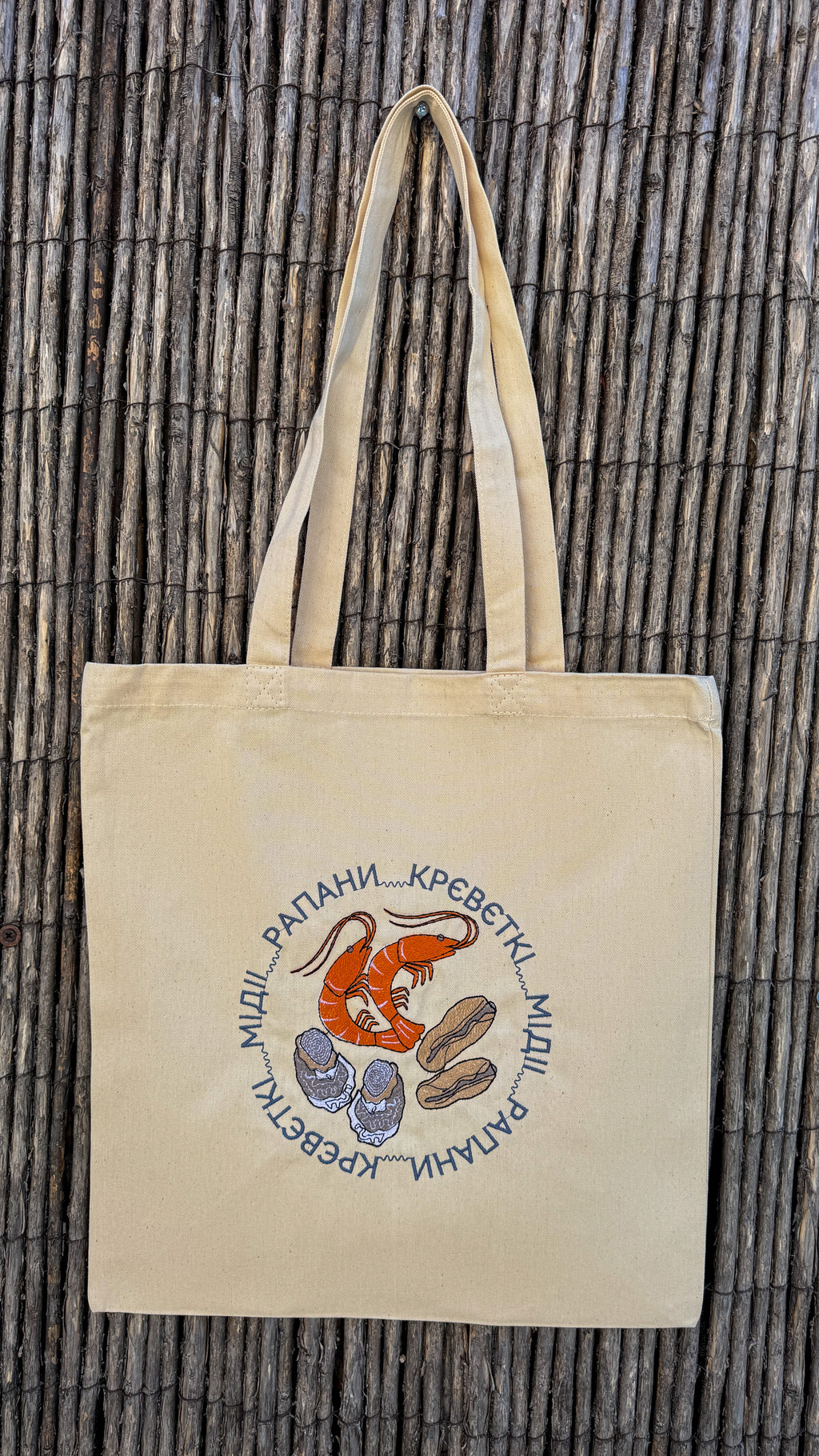 A beige canvas tote bag with two handles, featuring a circular graphic of a red shrimp and blue clams surrounded by Cyrillic text, displayed against a brown bamboo wall.