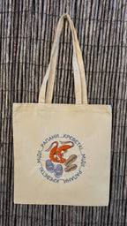 A beige canvas tote bag with two handles, featuring a circular graphic of a red shrimp and blue clams surrounded by Cyrillic text, displayed against a brown bamboo wall.