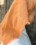 A close-up of a person wearing a soft, lightweight terracotta orange scarf or shawl draped over their shoulder and neck, paired with light blue denim jeans against a dark textured background.