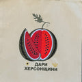 A beige canvas tote bag with a embroidered watermelon graphic and Cyrillic text, hanging against a dark brown woven bamboo background.
