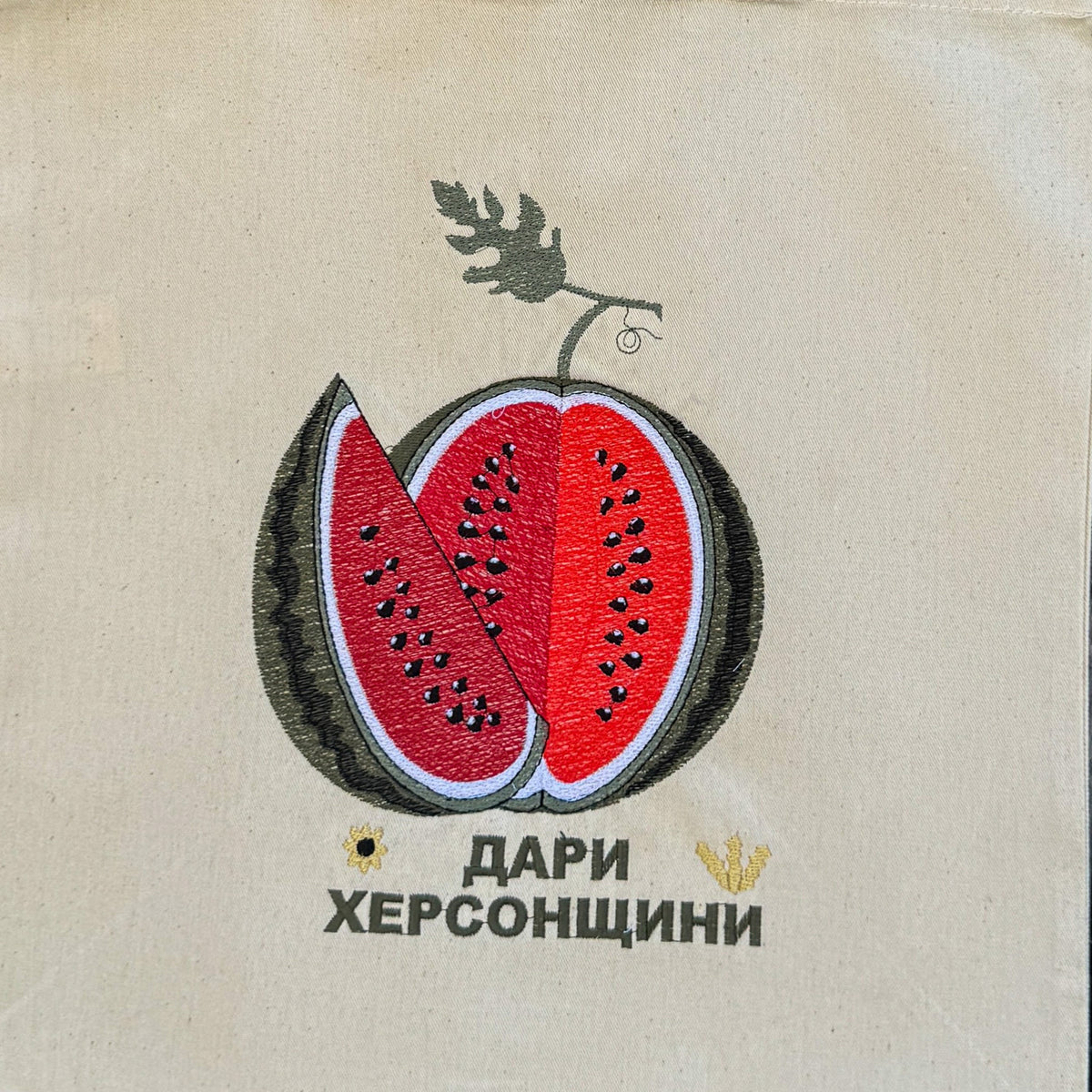 A beige canvas tote bag with a embroidered watermelon graphic and Cyrillic text, hanging against a dark brown woven bamboo background.
