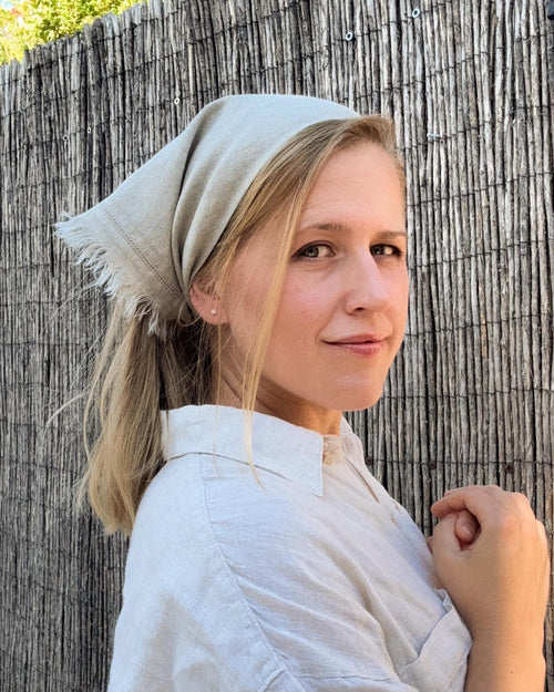 A woman wearing a light beige linen headscarf and matching linen shirt, posing in front of a rustic wooden fence.