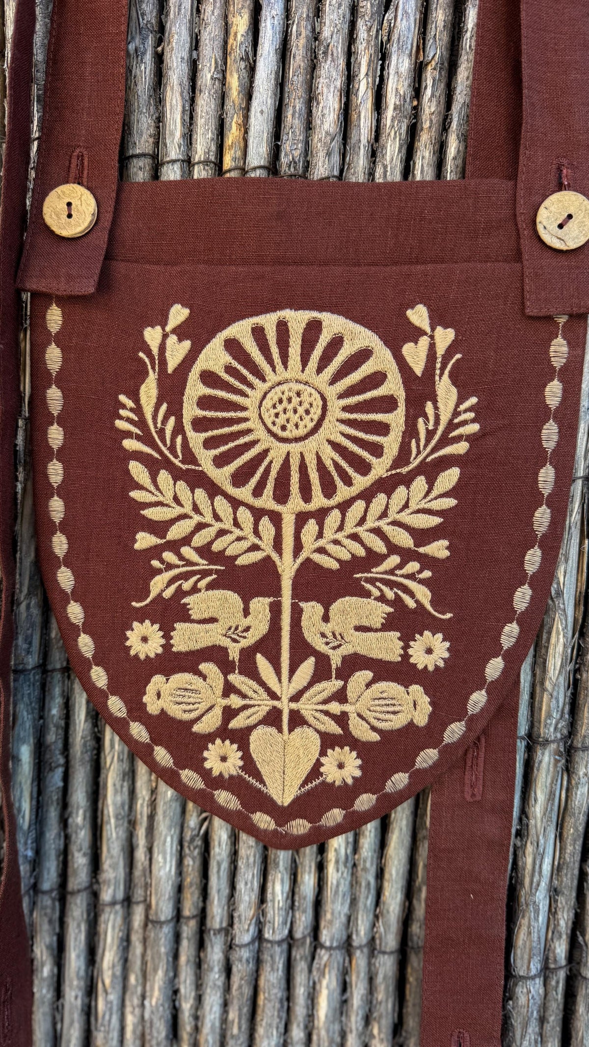 A close-up of a brown linen apron or pouch with a shield-shaped embroidered panel featuring a gold floral and bird motif, shown against a rustic bamboo background.
