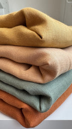 A stack of four folded textured fabric pieces in mustard yellow, tan, sage green, and rust orange, displayed on a white surface.