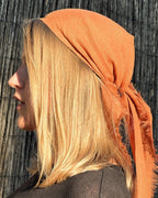 A side-profile view of a woman with blonde hair wearing an orange linen headscarf, set against a dark wooden background.