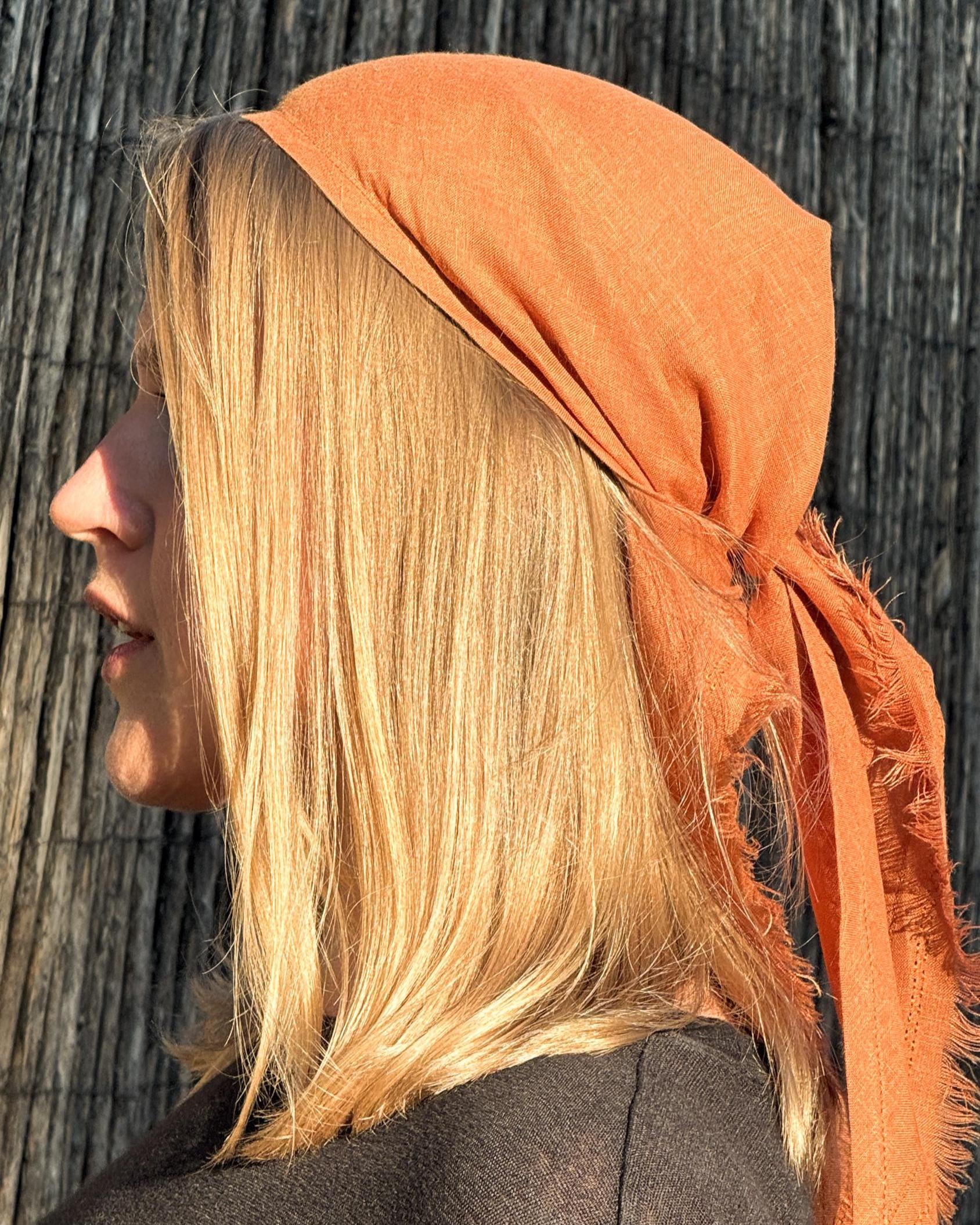 A side-profile view of a woman with blonde hair wearing an orange linen headscarf, set against a dark wooden background.