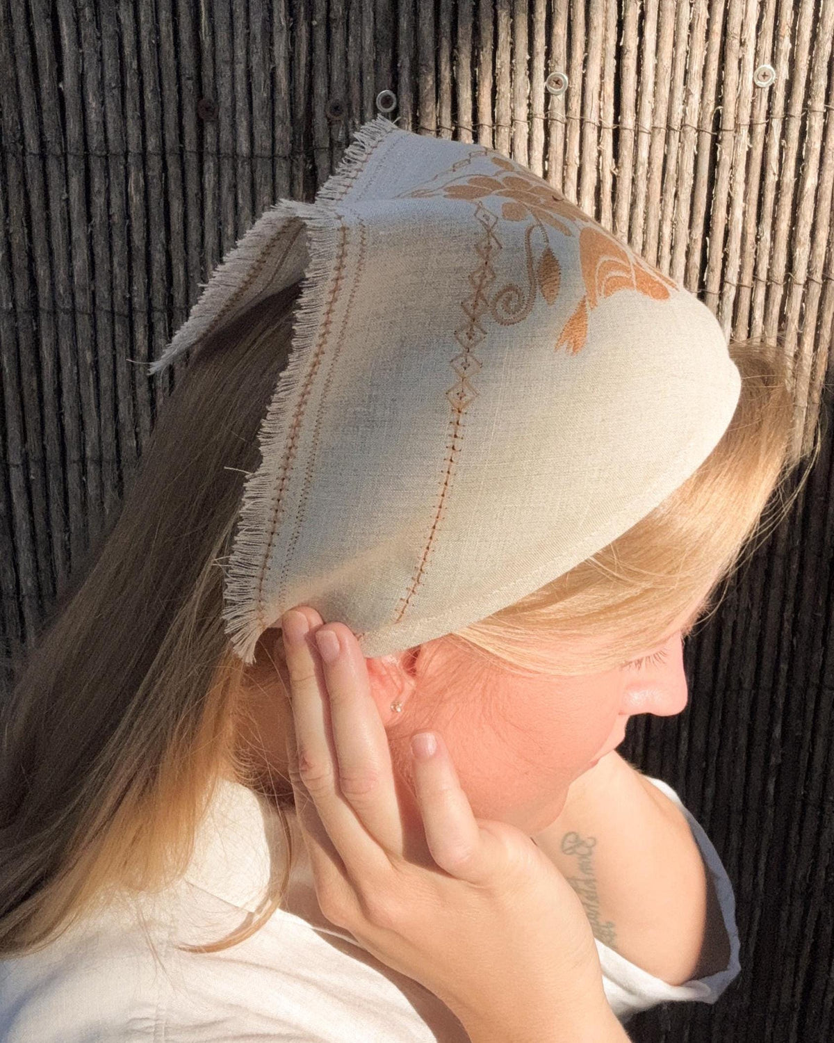 A woman wearing a light beige linen head covering with orange embroidered details and fringed edges, shown in profile against a dark bamboo wall.