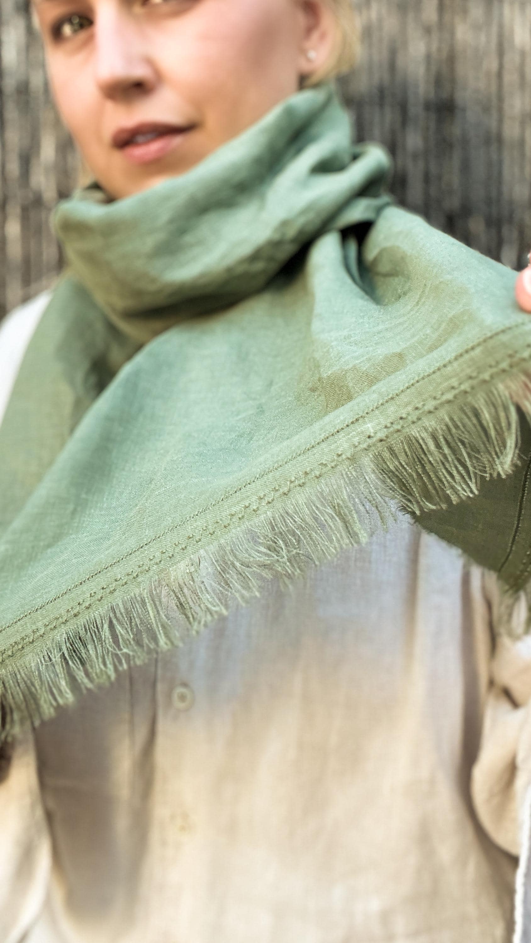 A close-up of a person wearing a sage green linen scarf with fringed edges, draped around their neck over a light beige linen shirt against a rustic wooden background.