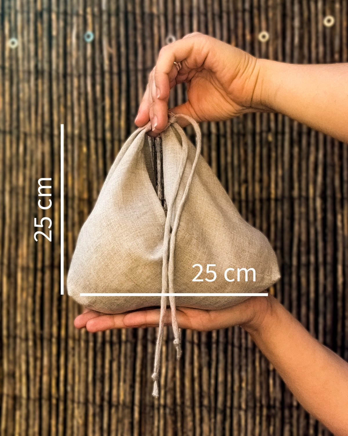 A small natural beige fabric drawstring pouch held in two hands against a dark woven bamboo background, with white measurement lines and text indicating 25 cm height and 25 cm width.