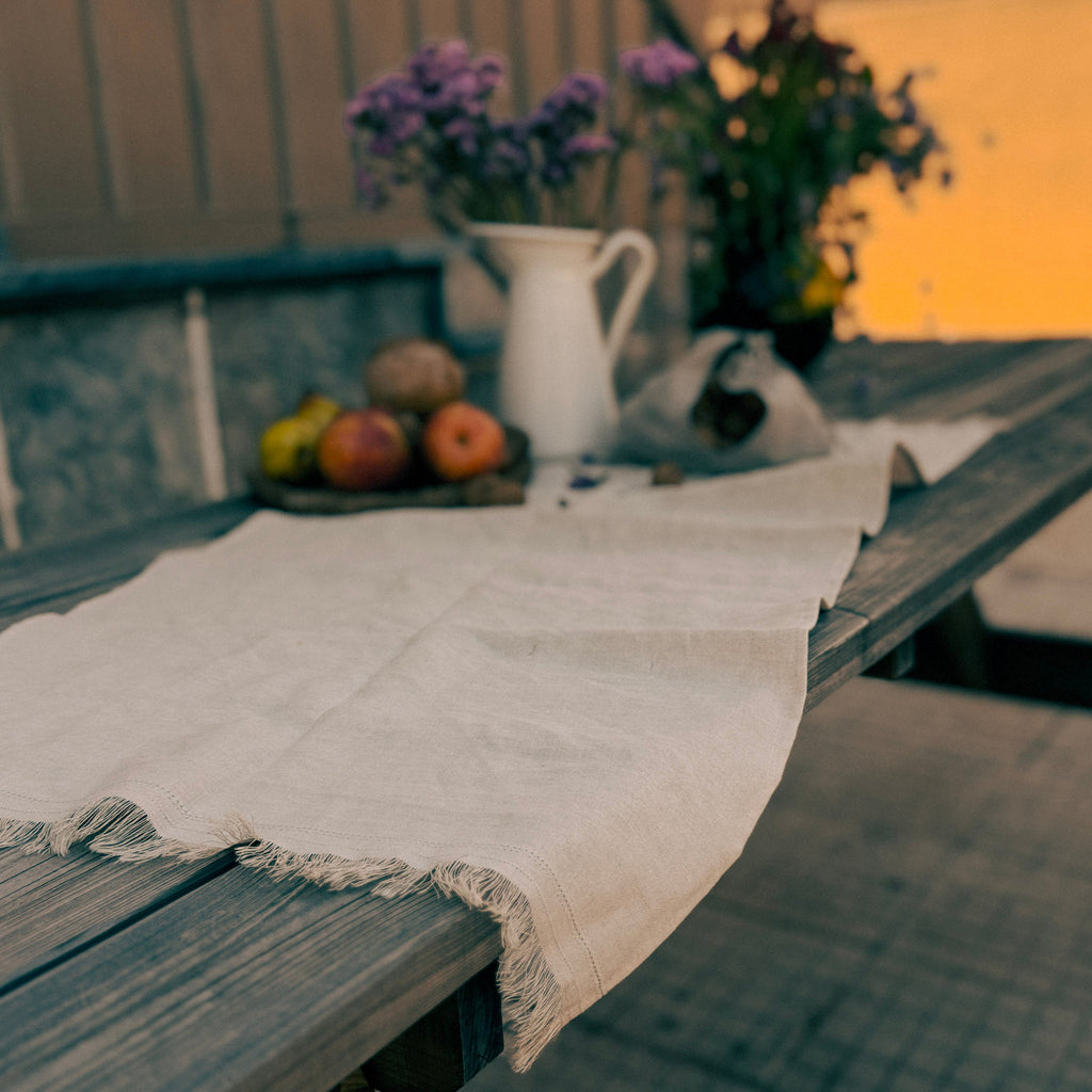 A rustic outdoor table setting with a draped off-white linen table runner, a white pitcher, and flowers in the background on a wooden deck.