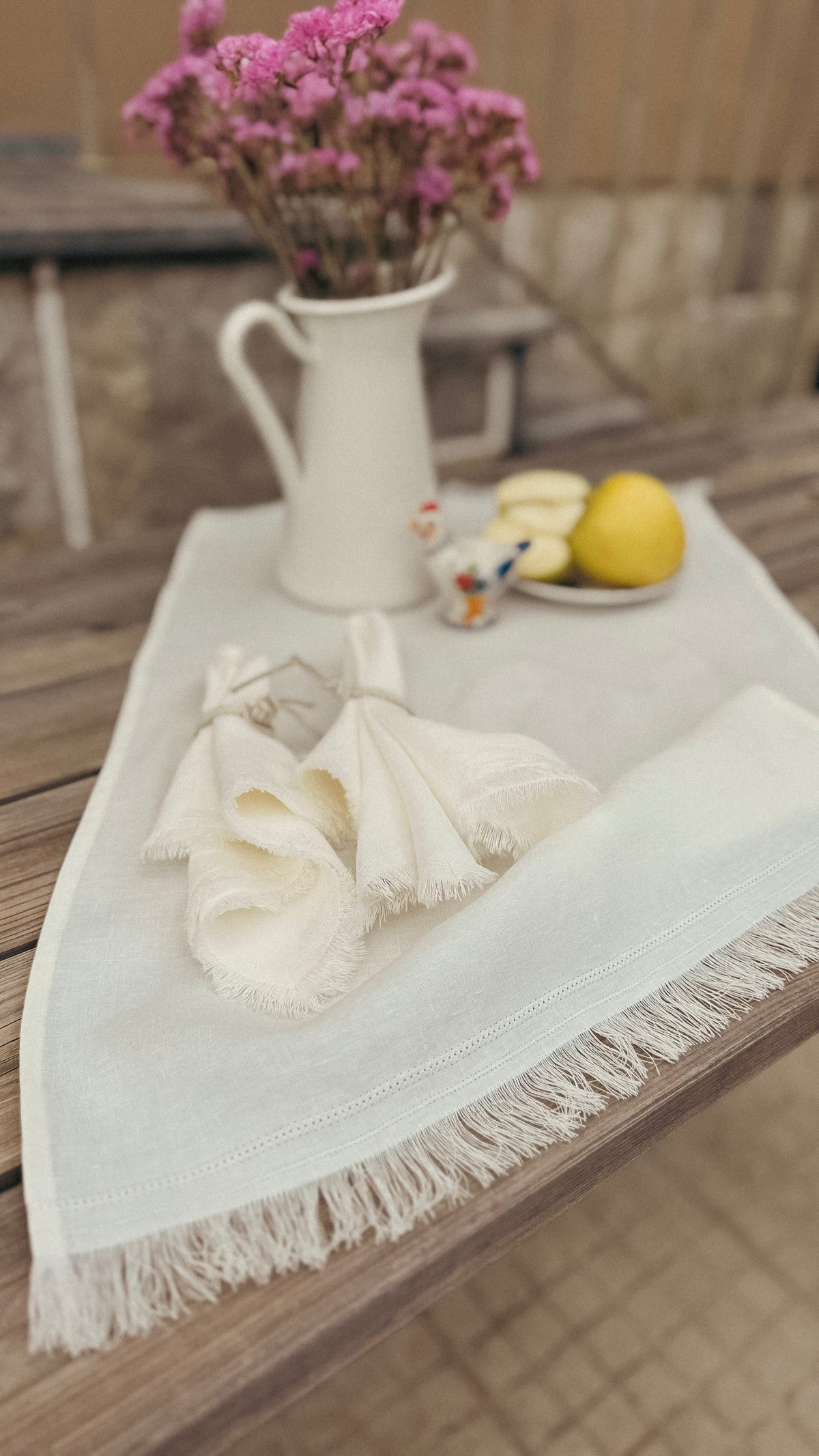 A cream-colored cotton table runner with fringe trim, styled on a wooden table with a matching napkin, a white pitcher of pink flowers, and a small plate with cheese and a lemon.