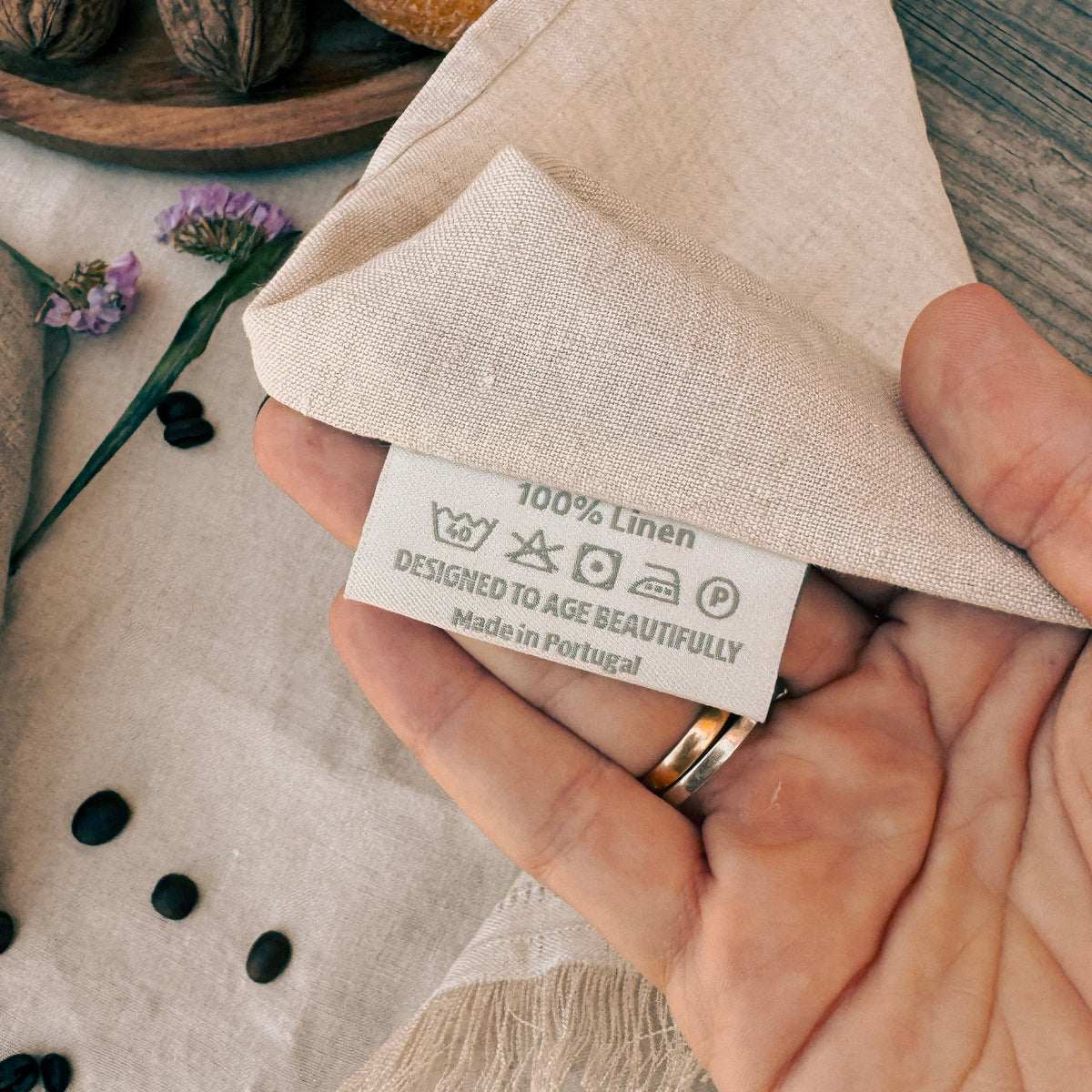 A close-up of a hand holding a folded beige linen fabric tag with a sewn-in label that reads '100% Linen' and 'Designed to Age Beautifully Made in Portugal.' The hand wears two rings, and the background includes dried flowers, dark beans, and a wooden surface.