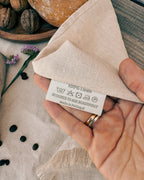 A close-up of a hand holding a folded beige linen napkin with a white fabric care label that reads '100% Linen' and 'Designed to Age Beautifully Made in Portugal.' The scene includes rustic bread, walnuts, and small flowers on a wooden surface, making this a lifestyle product shot.