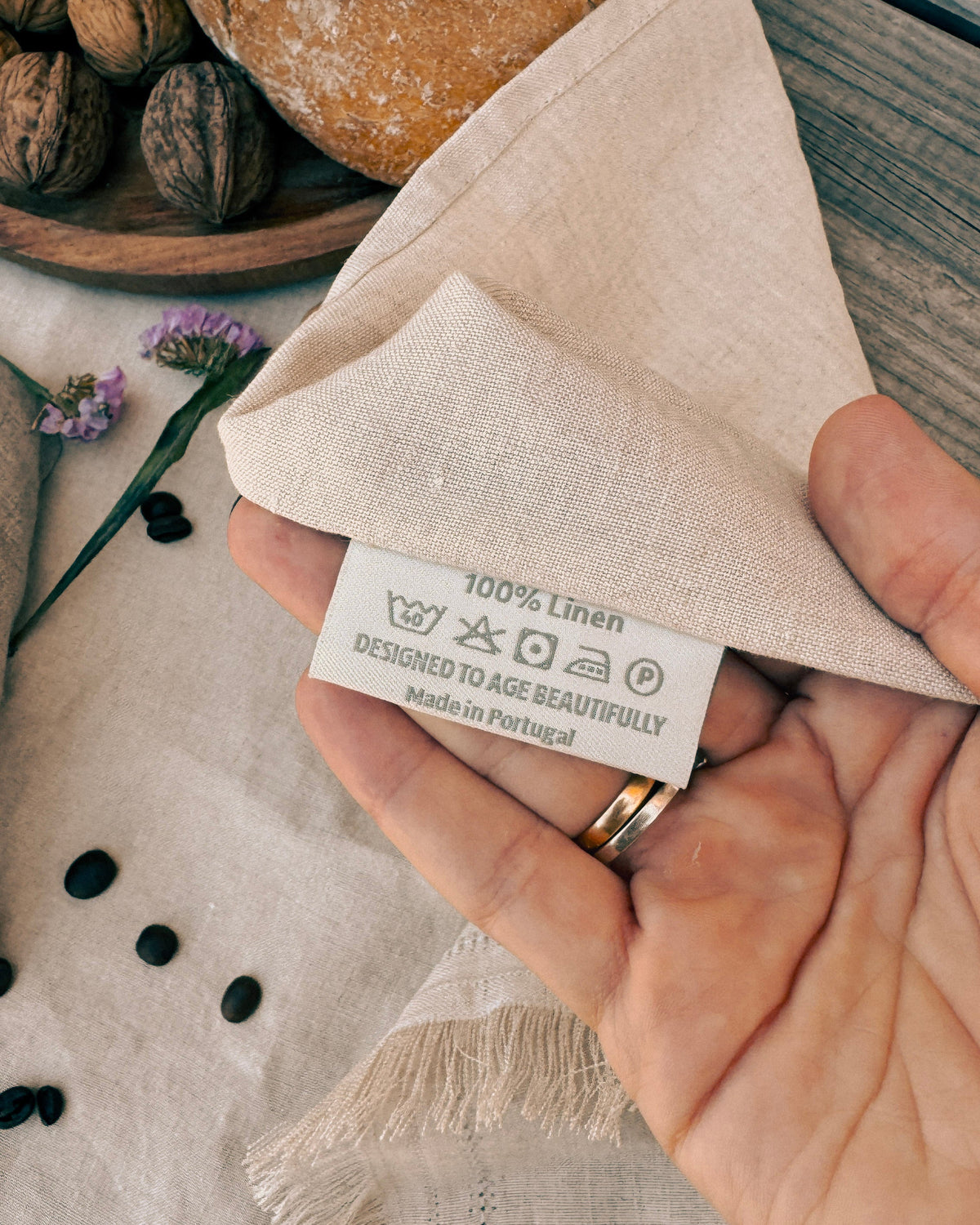 A close-up of a hand holding a folded beige linen napkin with a white fabric care label that reads '100% Linen' and 'Designed to Age Beautifully Made in Portugal.' The scene includes rustic bread, walnuts, and small flowers on a wooden surface, making this a lifestyle product shot.