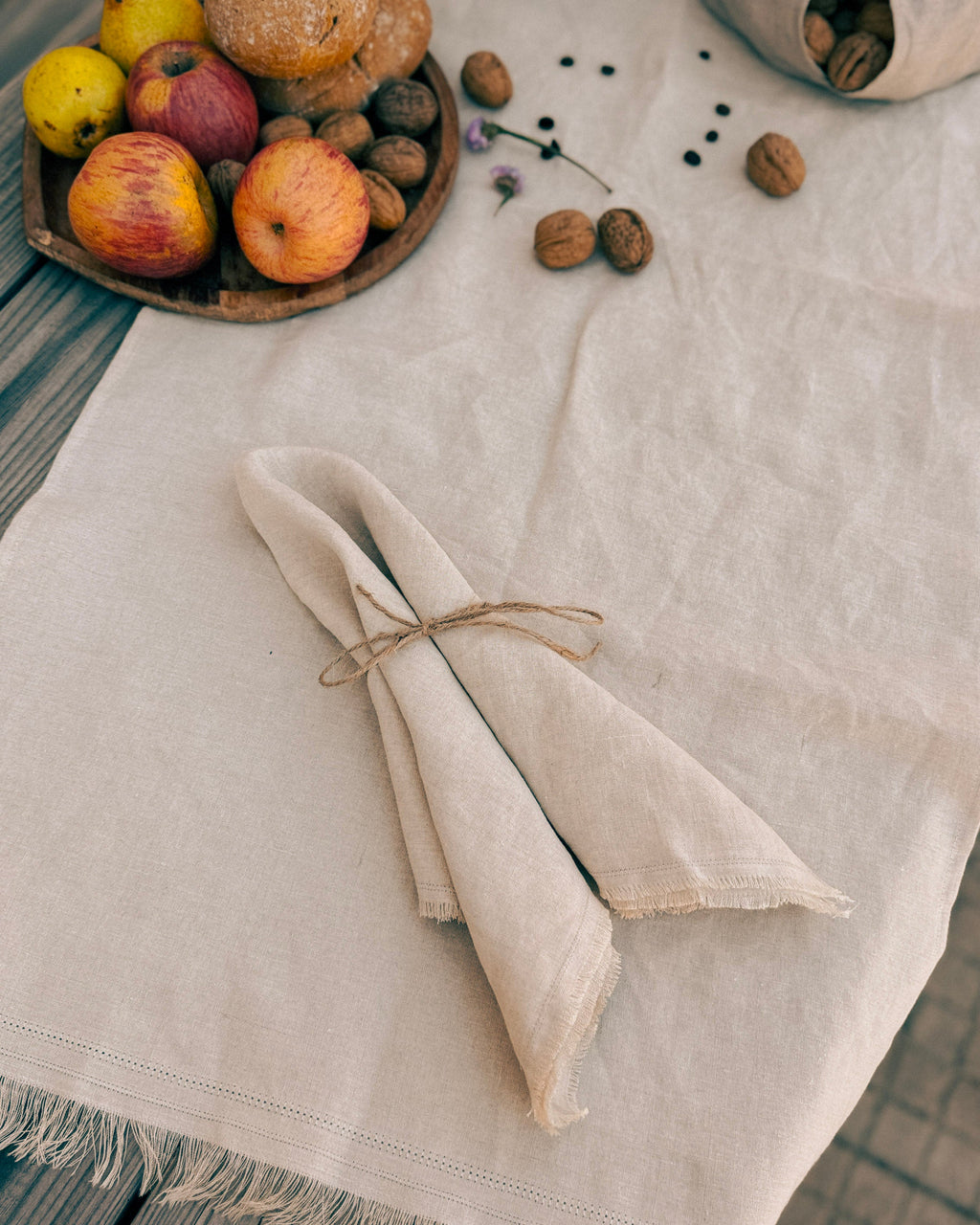A folded natural linen napkin tied with twine sits on a light beige linen tablecloth, styled with apples, walnuts, and a wooden bowl on a rustic wooden table.