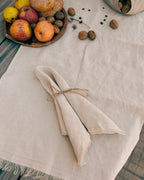 A folded natural linen napkin tied with twine sits on a light beige linen tablecloth, styled with apples, walnuts, and a wooden bowl on a rustic wooden table.