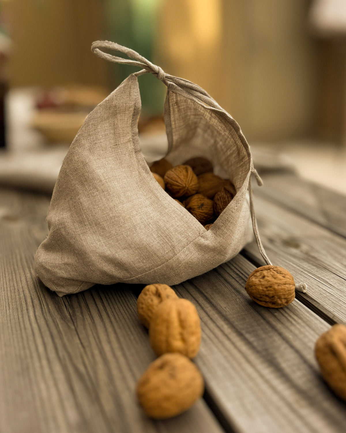 A small beige linen drawstring bag spilling walnuts onto a rustic wooden table, with a softly blurred outdoor background.