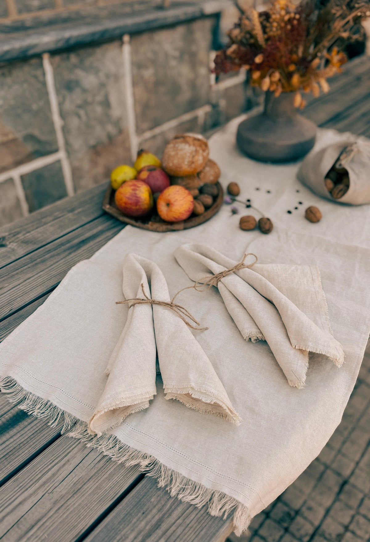 Two folded off-white linen napkins tied with twine are arranged on a rustic wooden table with a matching fringed runner. A small tray of fruit, bread, nuts, and a vase of dried flowers sit in the background.