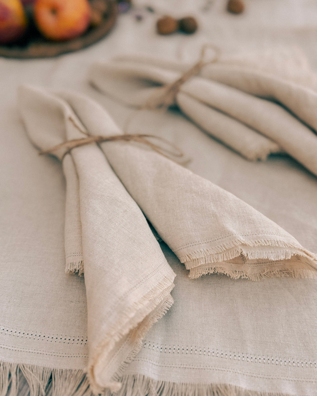 Natural linen table linens, including folded napkins tied with twine and a matching fringed tablecloth, styled on a rustic table with warm autumn props.