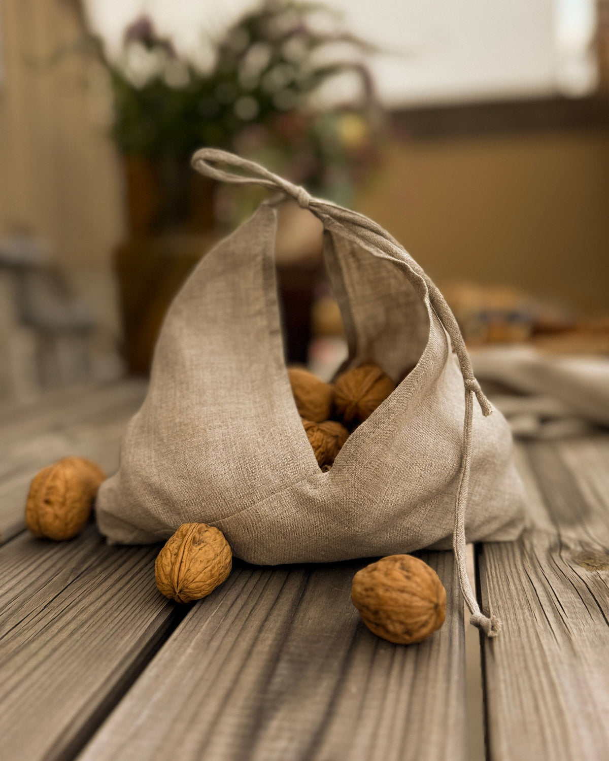 A small natural linen drawstring bag filled with walnuts, placed on a weathered wooden surface with a few walnuts scattered around it. The background is softly blurred, showing an indoor setting with warm tones.