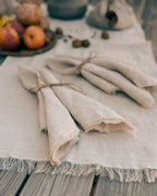Three folded beige linen napkins tied with twine, arranged on a matching fringed table runner over a rustic wooden table with apples and nuts in the background.
