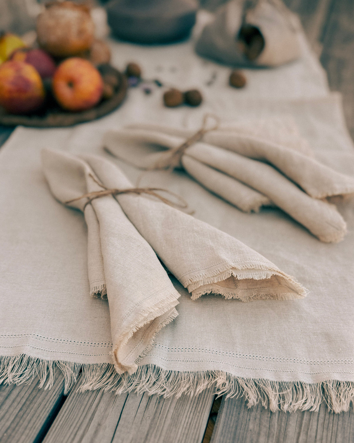 Three folded beige linen napkins tied with twine, arranged on a matching fringed table runner over a rustic wooden table with apples and nuts in the background.