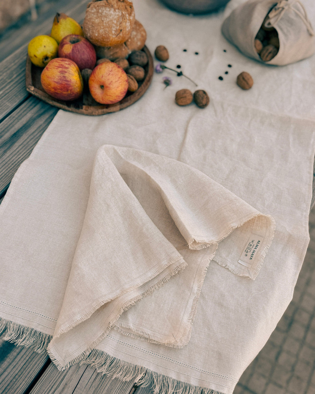 A light beige linen table runner with fringed edges draped over a weathered gray wooden table, styled with a small wooden tray of apples, bread, walnuts, and a matching fabric bag.