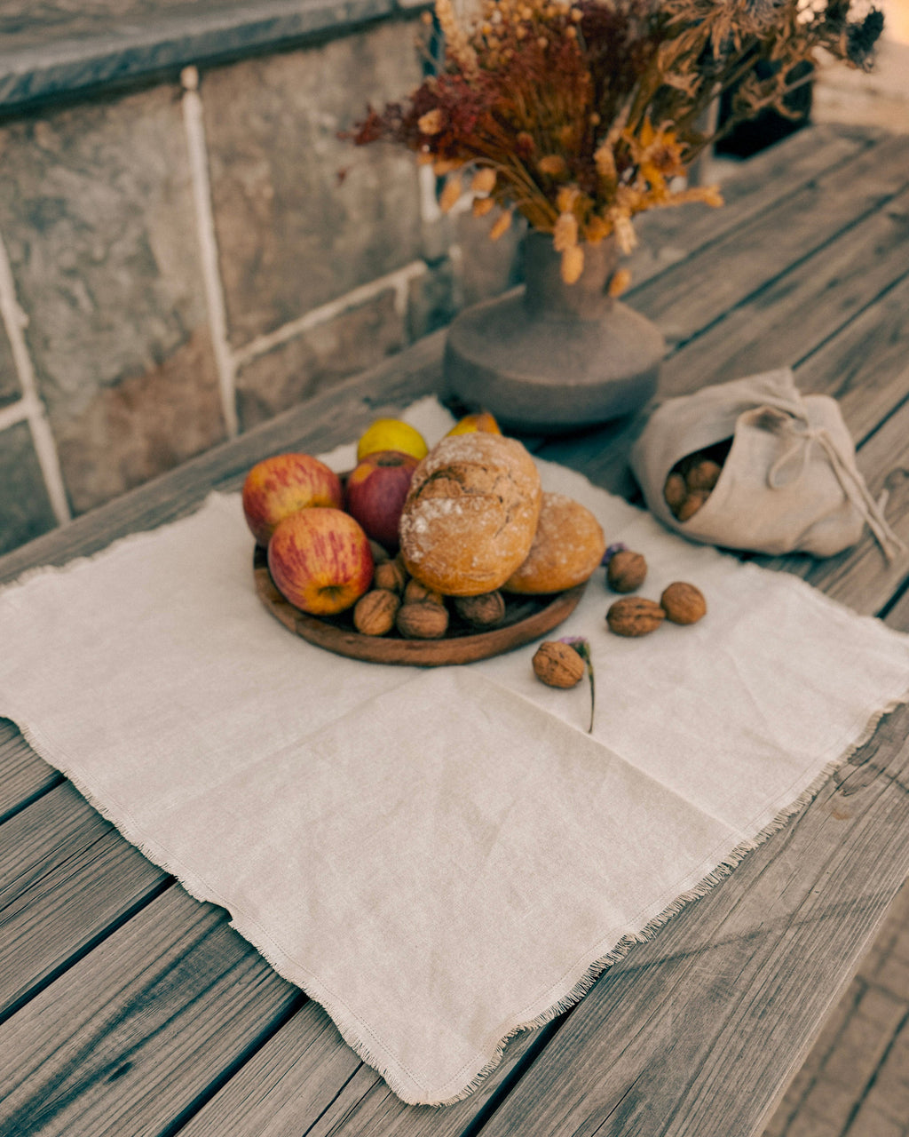 A rustic styled tabletop scene with a white linen cloth on weathered wooden planks, holding a small plate of bread, apples, and walnuts, with a vase of dried flowers and a drawstring bag in the background.