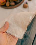 A close-up of a hand holding a corner of a light beige linen napkin with fringed edges, set against a rustic background with wooden textures and blurred walnuts.