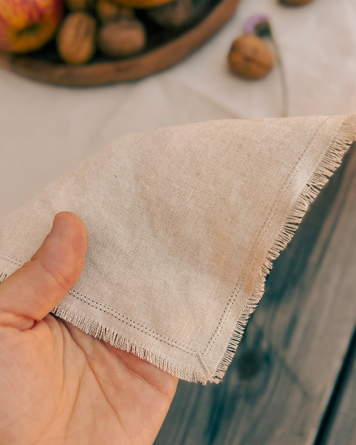 A close-up of a hand holding a corner of a light beige linen napkin with fringed edges, set against a rustic background with wooden textures and blurred walnuts.