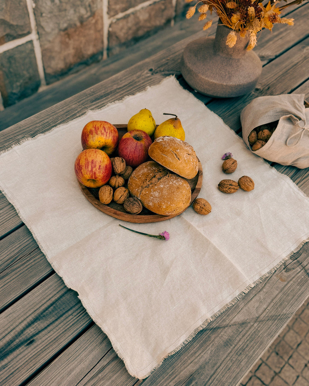 A styled flat-lay of rustic bread rolls and apples on a wooden board, set on a cream linen cloth on a weathered wooden table. A small vase with dried flowers and a beige drawstring bag of walnuts are nearby.