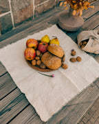 A styled flat-lay of rustic bread rolls and apples on a wooden board, set on a cream linen cloth on a weathered wooden table. A small vase with dried flowers and a beige drawstring bag of walnuts are nearby.