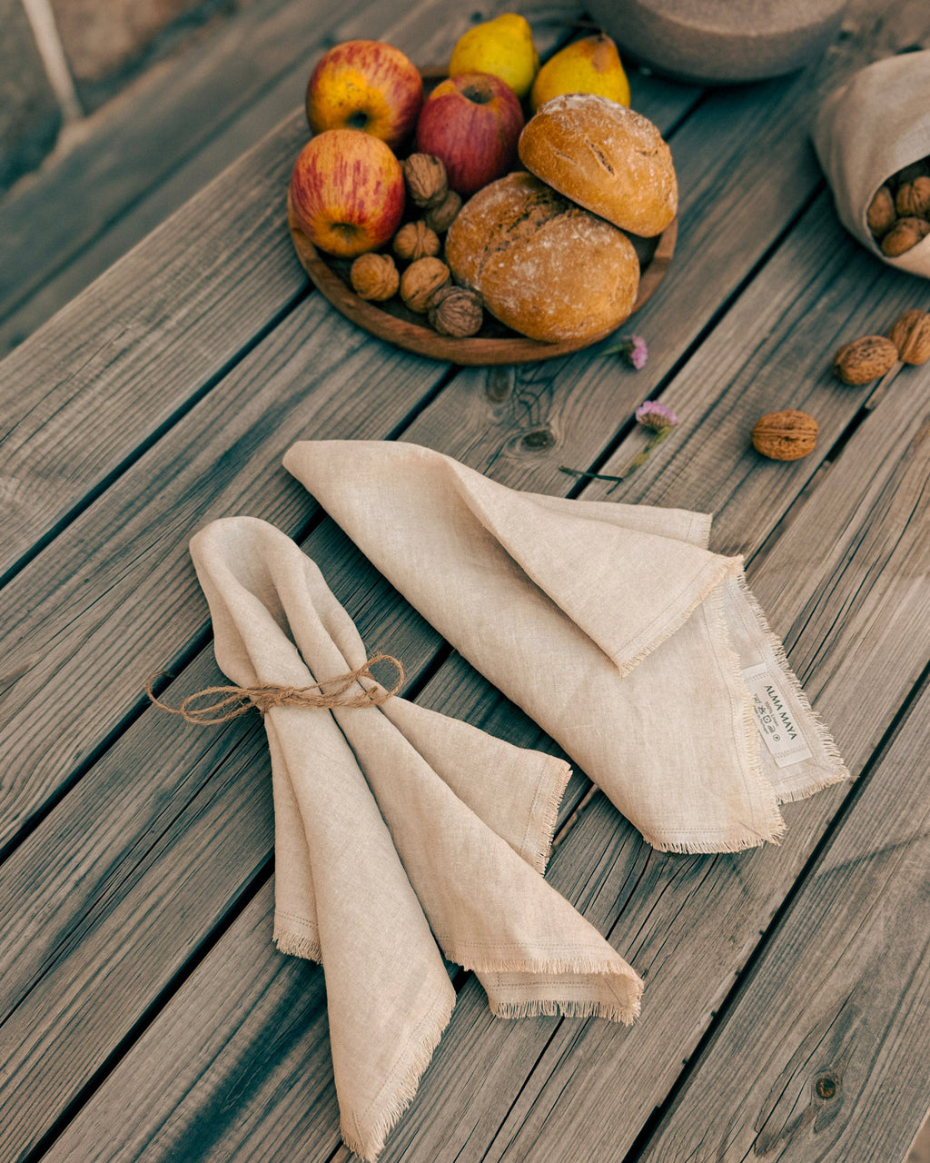 Two folded natural linen napkins tied with twine and laid on a weathered wooden table, with a bowl of bread, apples, and walnuts in the background.