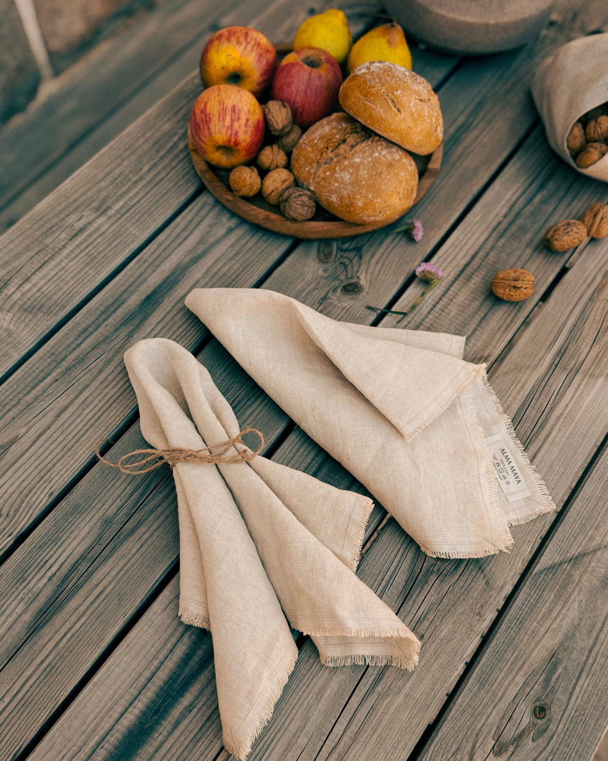 Two folded natural linen napkins tied with twine and laid on a weathered wooden table, with a bowl of bread, apples, and walnuts in the background.