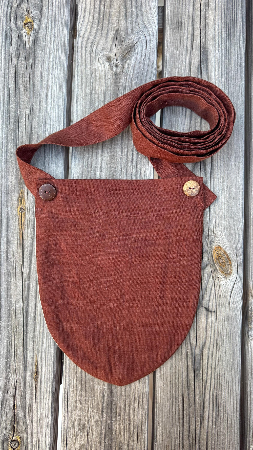 A rust-brown linen shoulder bag with a matching adjustable strap, laid flat on a weathered gray wooden plank background.