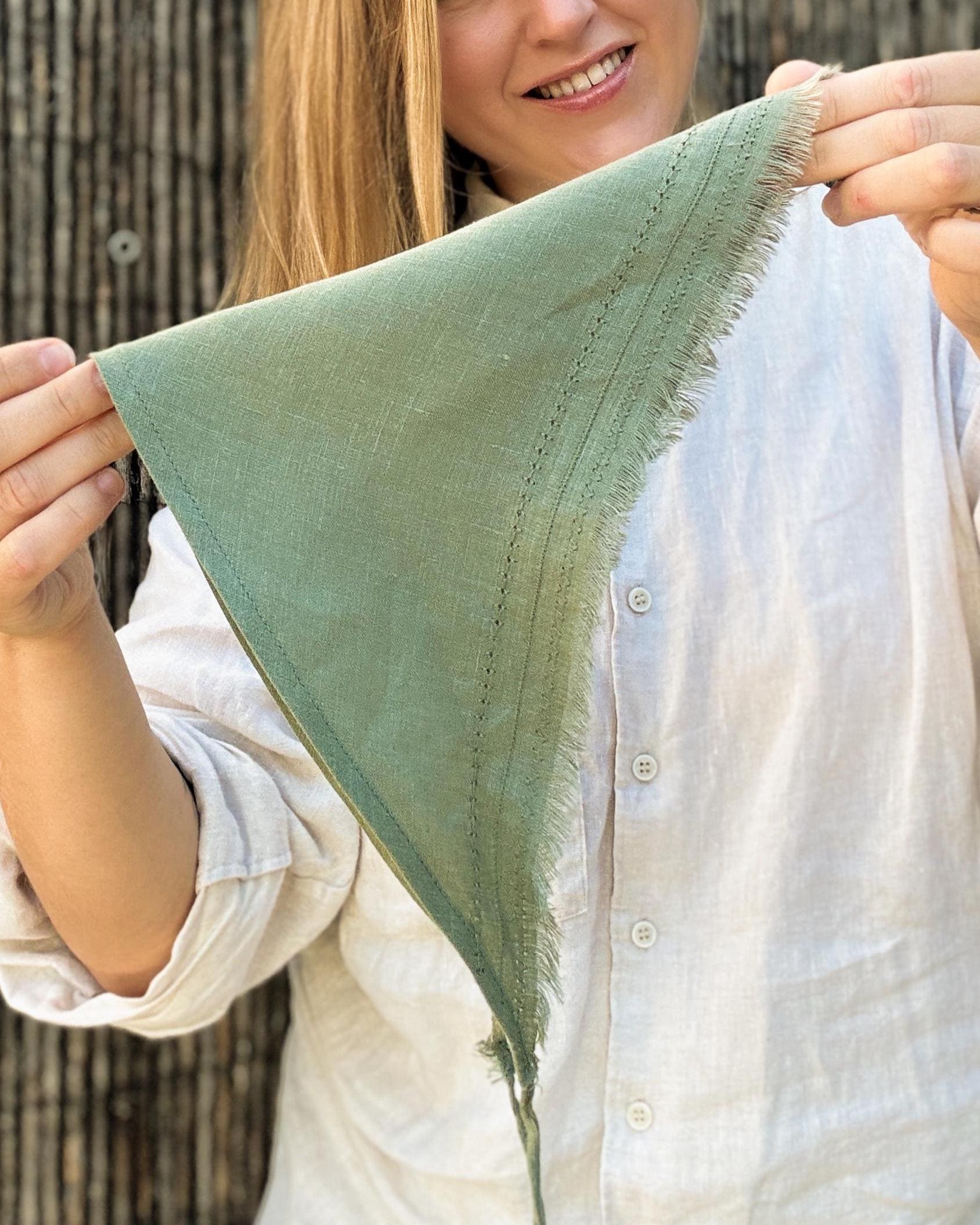 A woman holding up a sage green triangular fringed bandana or scarf, wearing a white linen button-up shirt against a rustic wooden background.