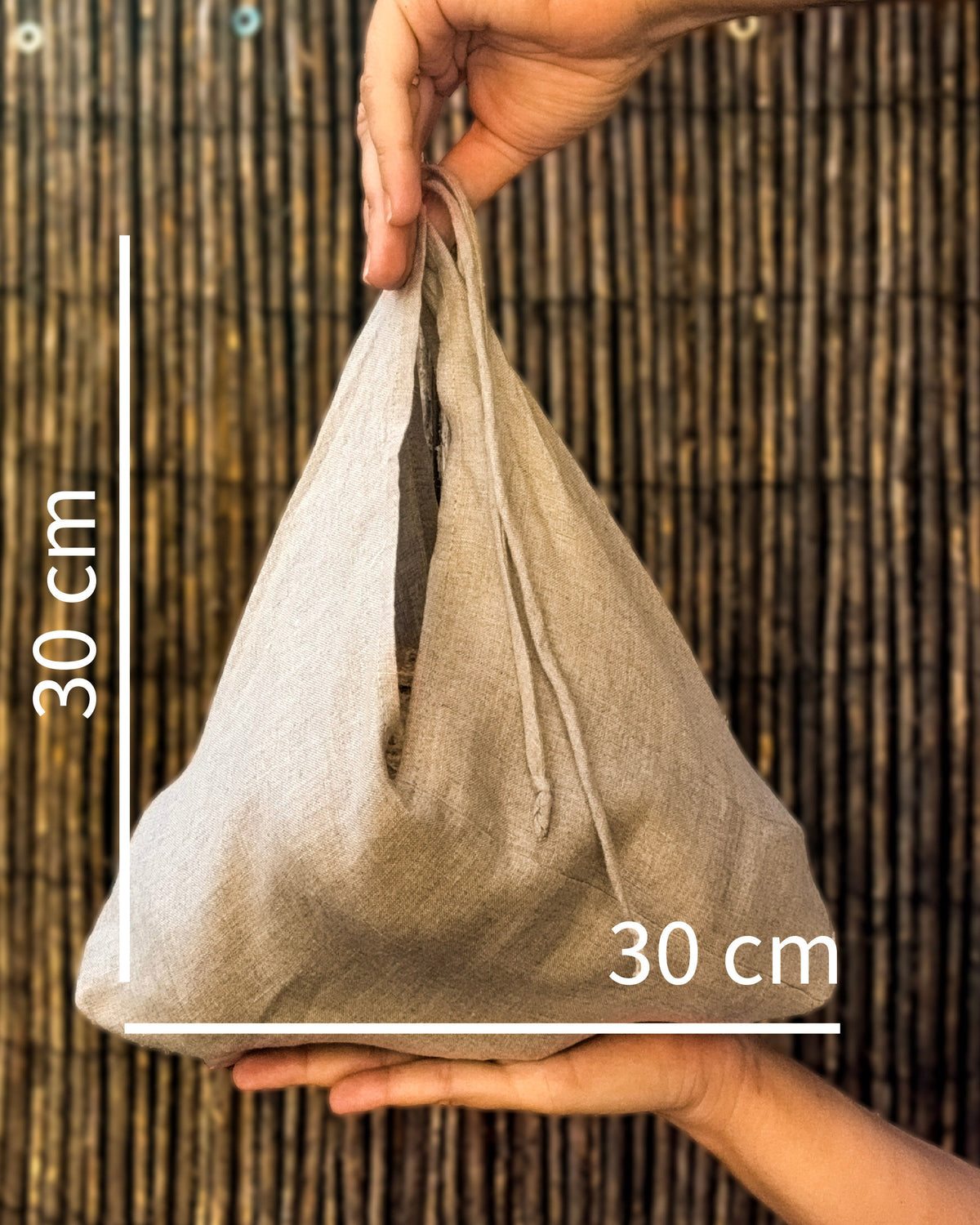 A small natural beige fabric drawstring pouch held in two hands against a dark woven bamboo background, with white measurement lines and text indicating 30 cm height and 30 cm width.