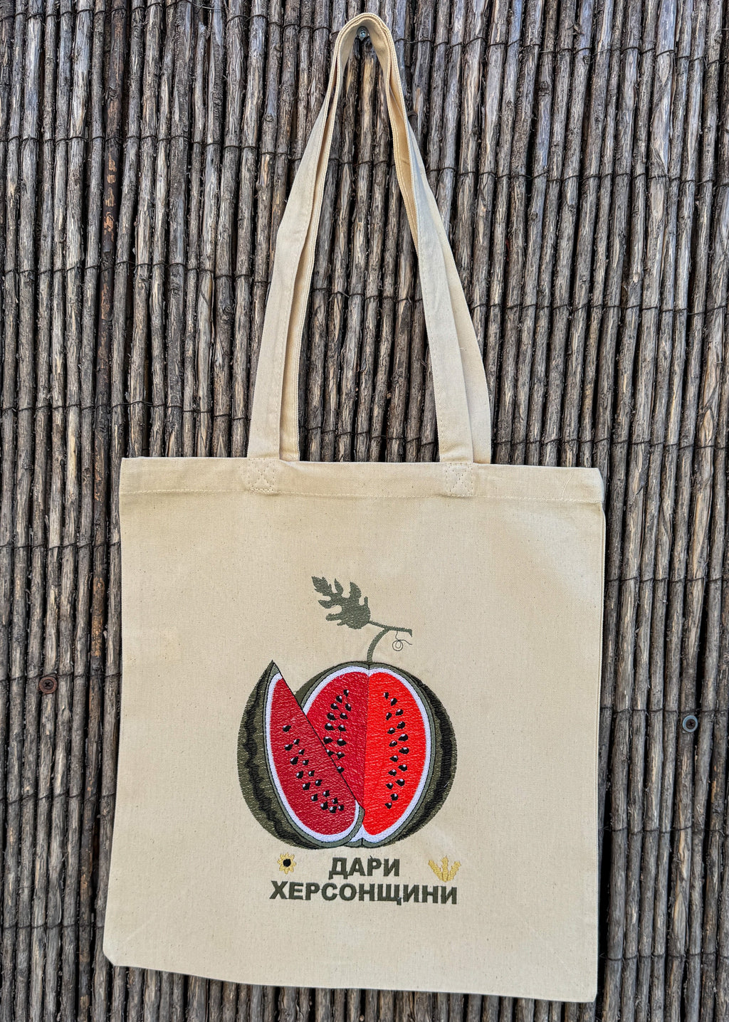 A natural canvas tote bag with a embroidered watermelon graphic and the Ukrainian text 'Дари Херсонщини' on the front, hanging against a vertical bamboo reed background.