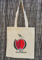 A natural canvas tote bag with a embroidered watermelon graphic and the Ukrainian text 'Дари Херсонщини' on the front, hanging against a vertical bamboo reed background.