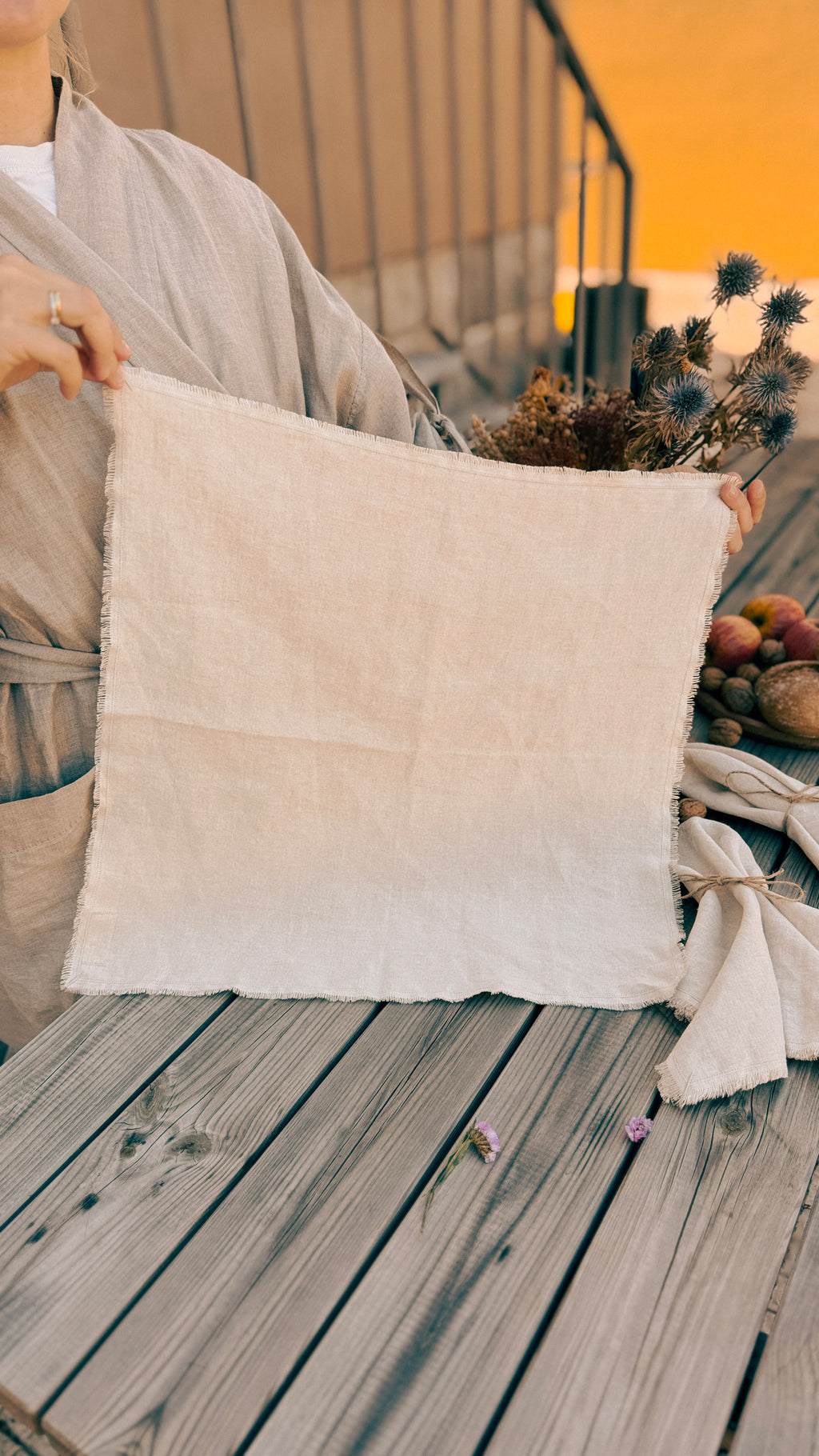 A person holds up a square, off-white linen fabric panel with raw, frayed edges on a weathered wooden deck. The person is wearing a light beige kimono-style robe, and dried flowers and a small basket of fruit sit on the deck in the background.