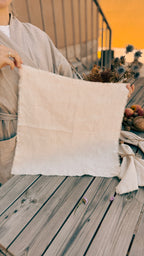 A person holds up a square, off-white linen fabric panel with raw, frayed edges on a weathered wooden deck. The person is wearing a light beige kimono-style robe, and dried flowers and a small basket of fruit sit on the deck in the background.
