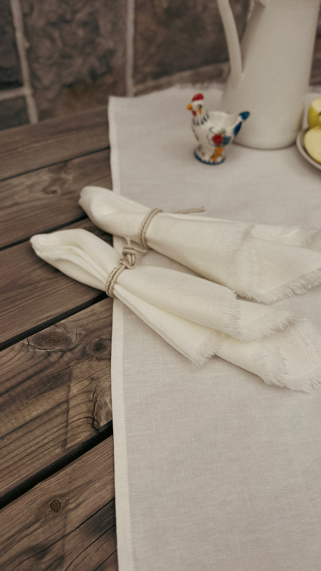 Two folded white napkins with fringed edges tied with twine, arranged on a light beige table runner over a rustic dark wood table. A small decorative rooster figurine and a white pitcher are visible in the background.
