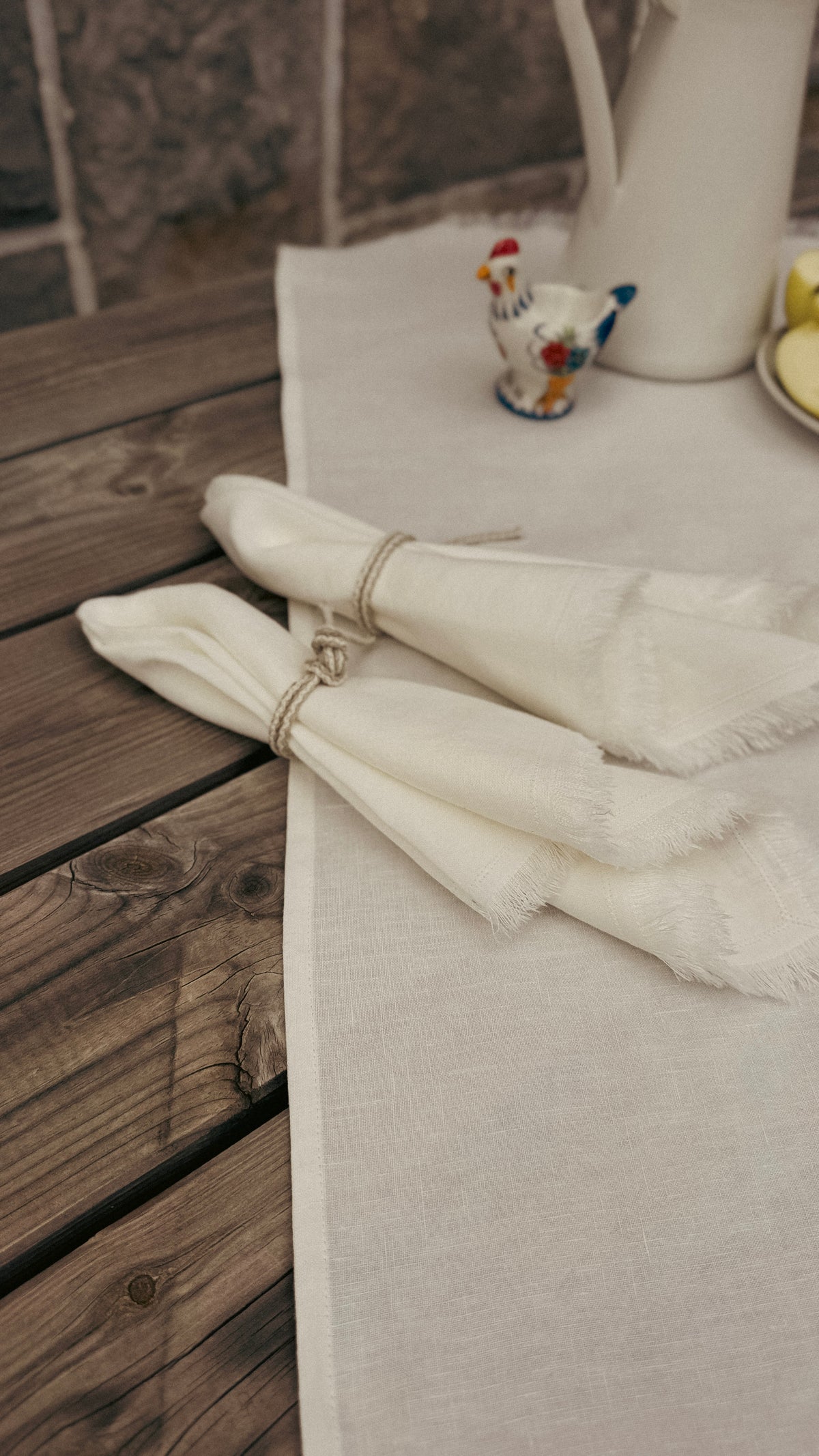 Two folded white napkins with fringed edges tied with twine, arranged on a light beige table runner over a rustic dark wood table. A small decorative rooster figurine and a white pitcher are visible in the background.