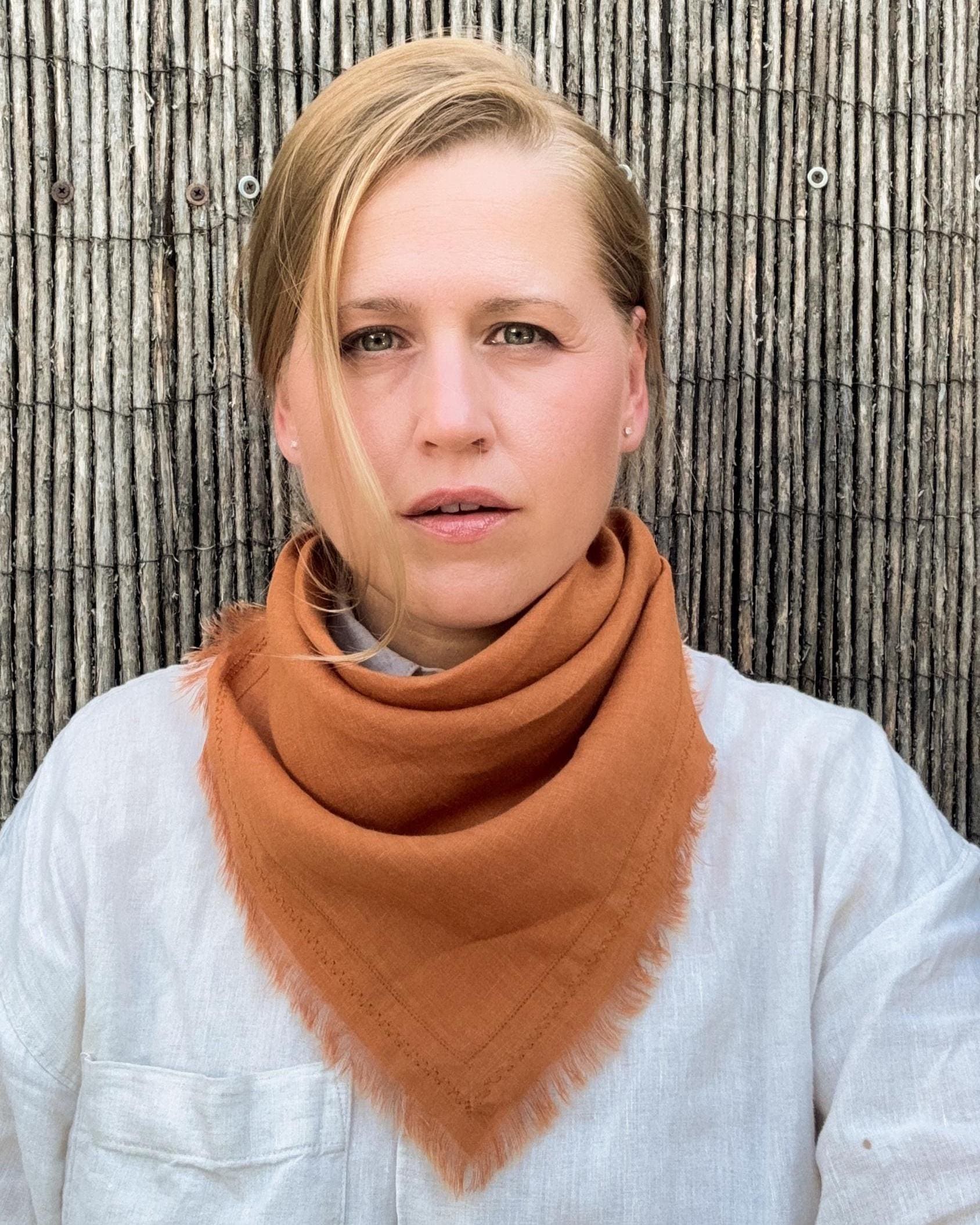 A woman wearing a warm rust-orange triangular bandana scarf with frayed edges, styled over a white linen shirt, standing against a vertical bamboo fence.