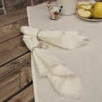 A rustic outdoor dining table styled with a white linen table runner, two folded white napkins tied with silver rings, a white ceramic pitcher with dried purple flowers, a small rooster figurine, and a plate of lemons.