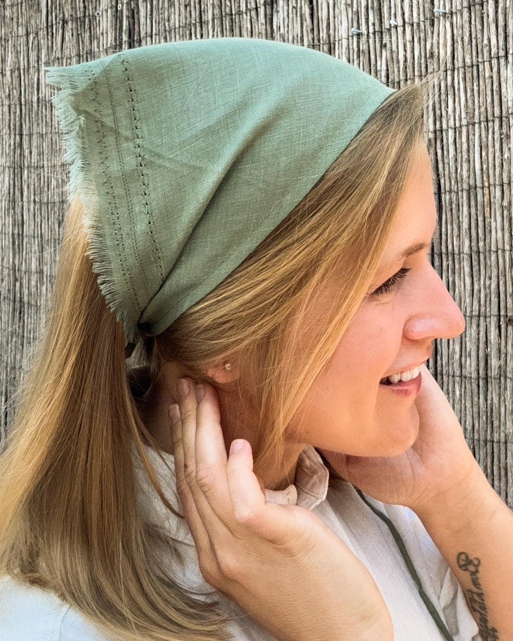 A woman wearing a sage green linen headscarf with frayed edges, shown in profile against a rustic bamboo wall.